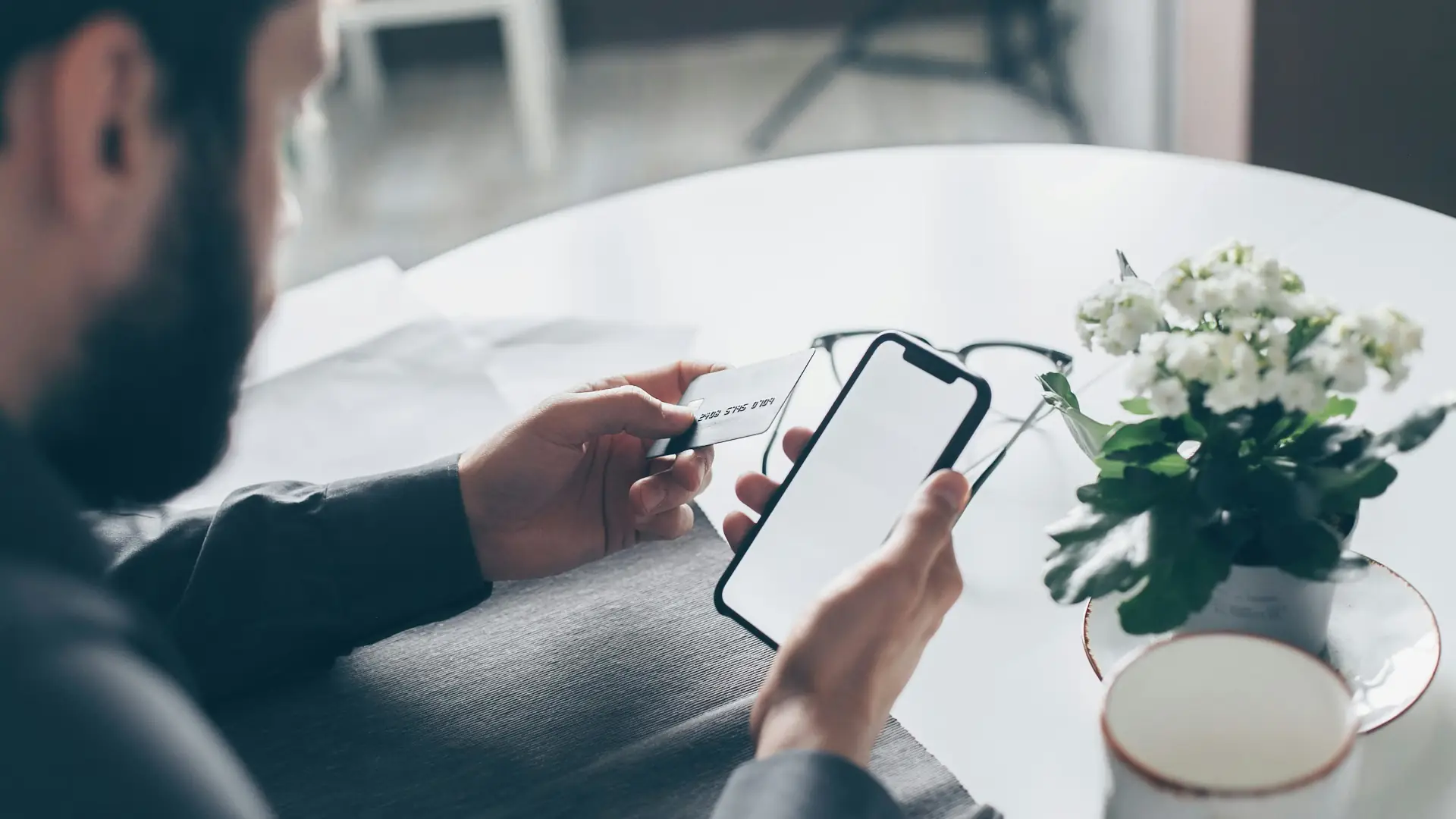 Henkilö tekee verkkokauppaostoksen puhelimella, luottokortti toisessa kädessä, valkoisen pöydän ääressä kukka ja silmälasit taustalla.