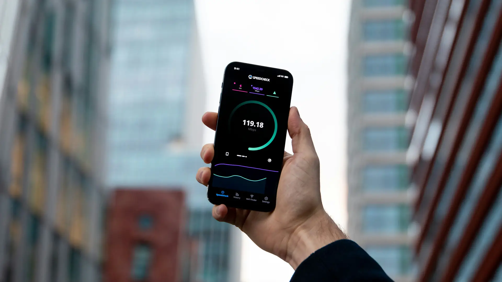 Käsi pitelee älypuhelinta, jonka näytöllä näkyy nopeustesti 119,18 Mbps, taustalla sumeat toimistotalot kaupungissa.