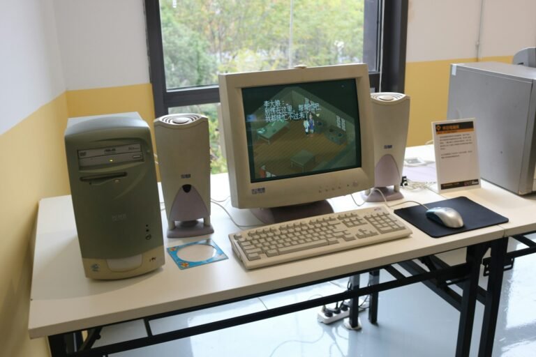 1990-luvun pöytäkone CRT-näytöllä, beige kotelo, näppäimistö, hiiri ja kaiuttimet valkoisella pöydällä ikkunan ääressä; hidas tietokone.
