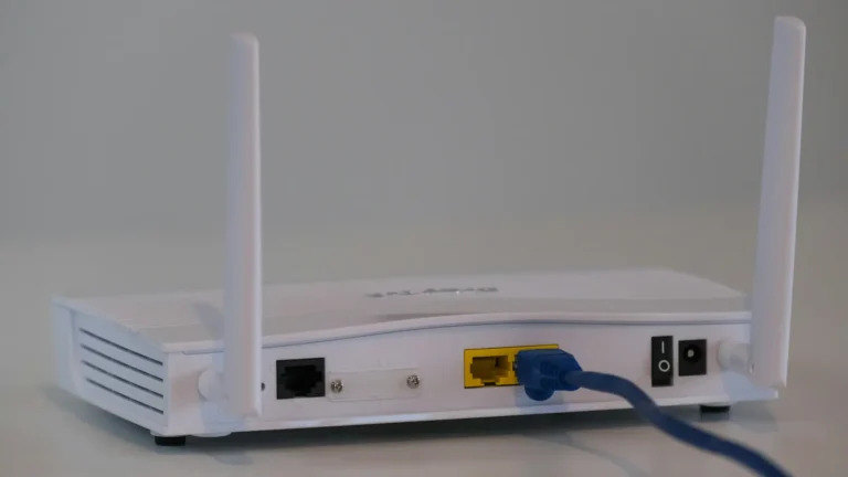 Valkoinen WiFi-reititin kahdella antennilla, takapaneelin portit näkyvissä ja sininen Ethernet-kaapeli kytkettynä, virtakytkin ja virtaliitäntä oikealla.